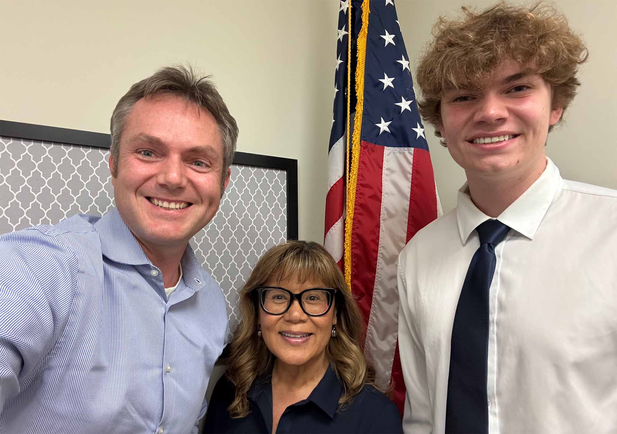 Three smiling people, woman with glasses between two men, US flag behind them.