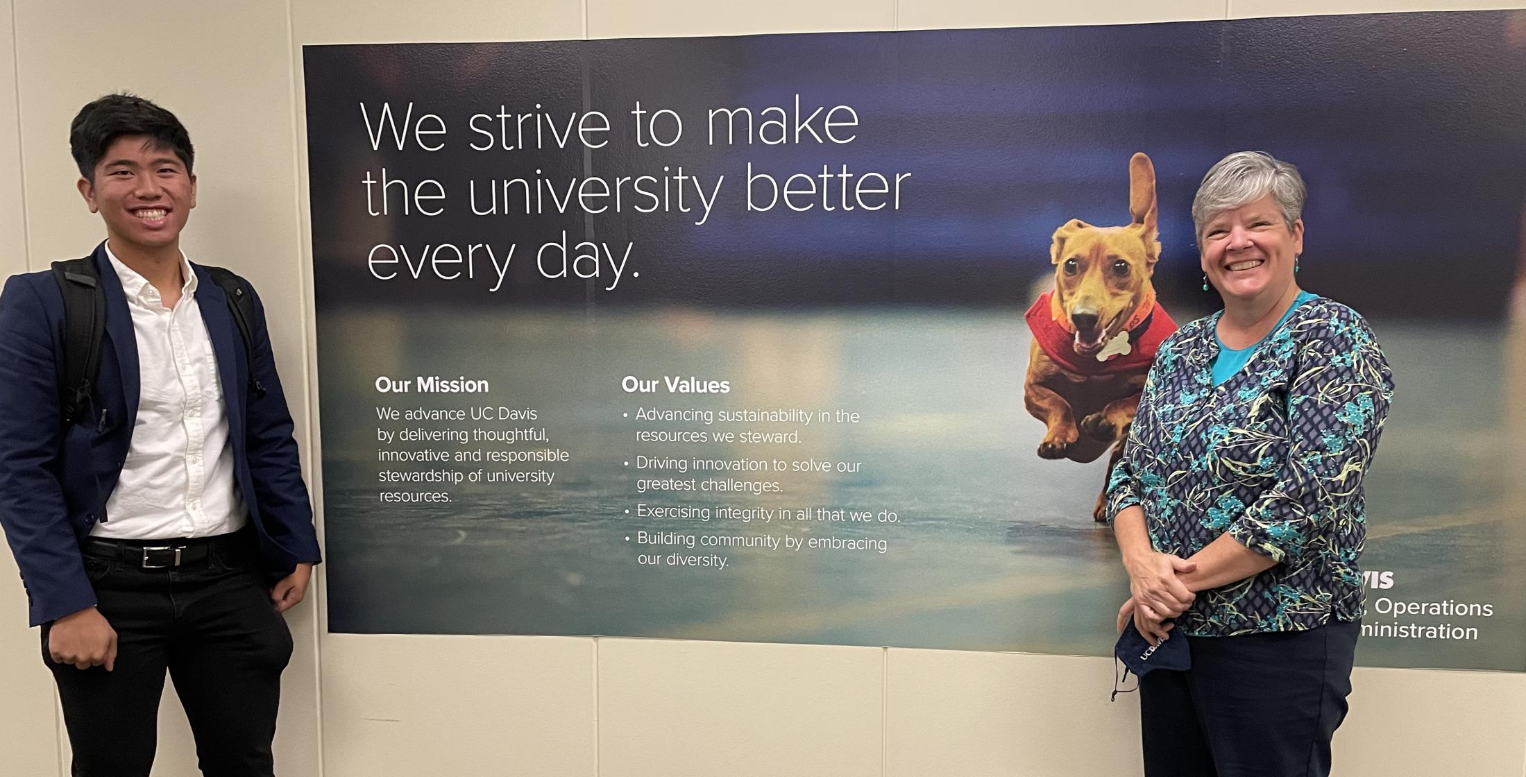 Two people stand in front of a sign in the Finance, Office and Administration office.