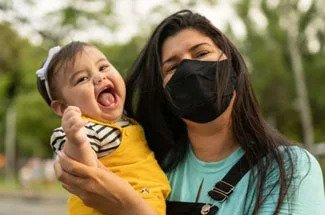 "Woman in face mask holds smiling infant"