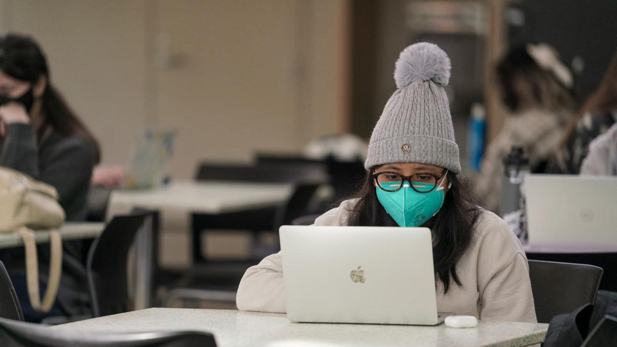 Woman, masked and wearing a knit cap, works at laptop