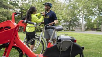 Maria Contreras Tebbutt, director of The Bike Campaign, adjusts UC Davis Chancellor Gary May's helmet strap as they stand outside. A red bike is in the foreground.
