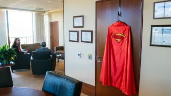 A Superman cape in the foreground and Chancellor May in the background chats with a student sitting down at UC Davis. 