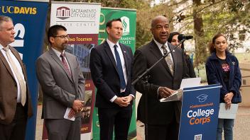 Photo of Chancellor Gary May speaking at a College Corps podium outside while people in suits stand behind him