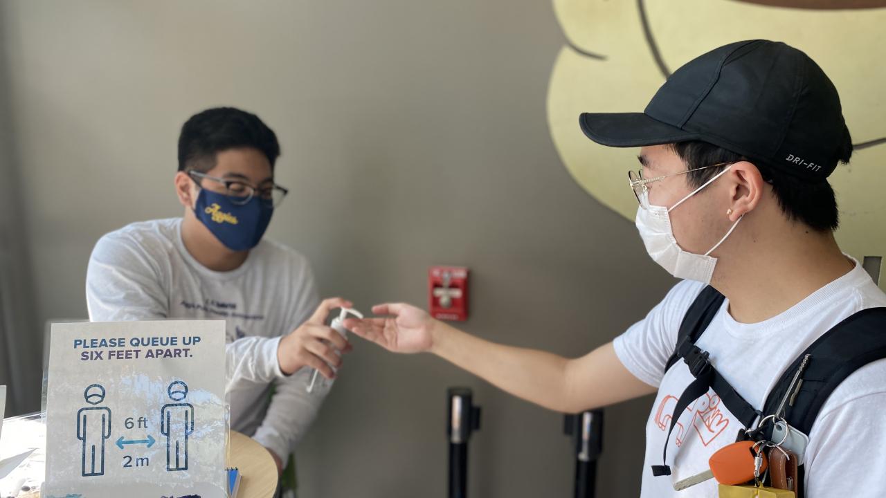 Masked public health ambassador providing hand sanitizer to student. 