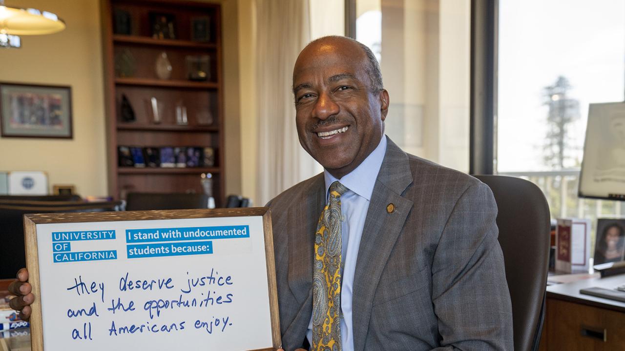 Chancellor Gary May looks into the camera smiling, holding a dry erase board that says, "I stand with undocumented students because they deserve justice and the opportunities all Americans enjoy".
