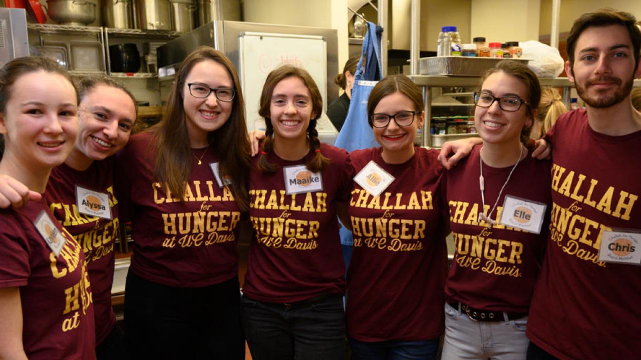 Students pose in a line in kitchen, all wearing burgundy T-shirts with “Challah for Hunger UC Davis” in gold.