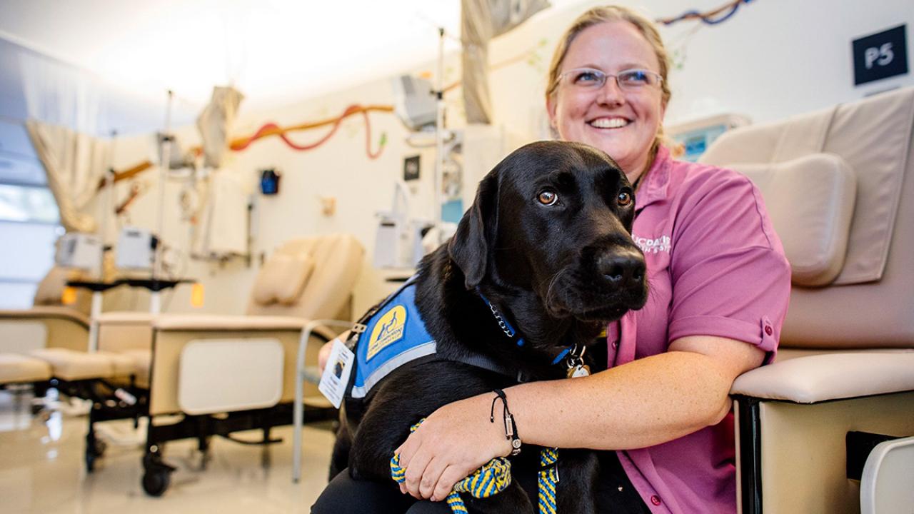 Jenny Belke sits in a medical office with Huggie, a black Labrador.