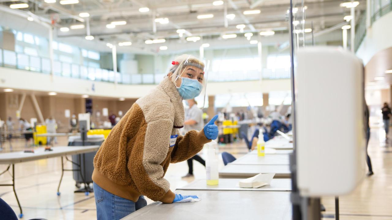 Aggie Public Health Ambassador wearing a mask, face shield and gloves wiping down a table and giving the thumbs up at COVID testing station.