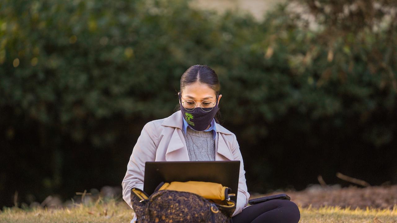 Student with face mask sits on ground while looking at an open laptop screen.