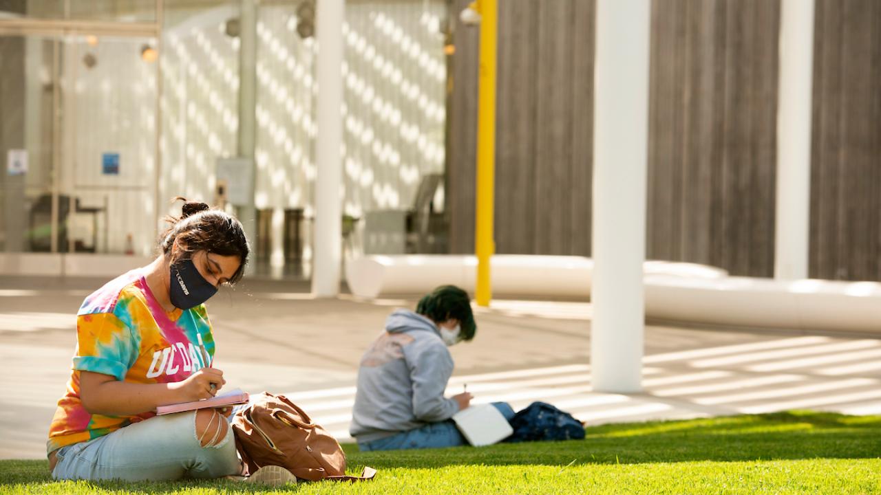 Two students wearing masks studying on lawn.