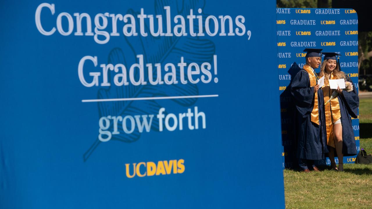 Sign that says "Congratulations, Graduates. Grow forth. UC Davis" Students in the background in caps and gowns.