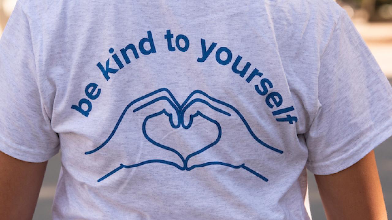 Grey t-shirt with blue text that reads "Be kind to yourself" with heart hands.