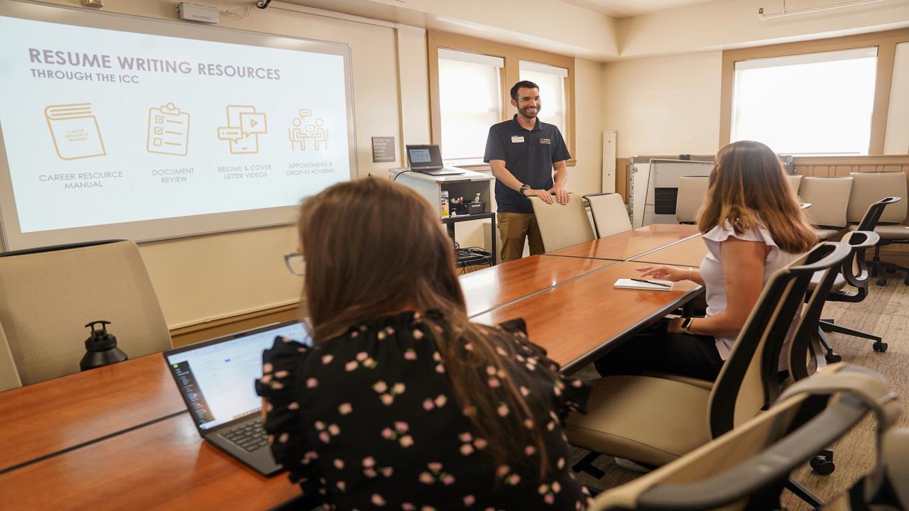 A male instructor from the UC Davis Internship and Career Center stands in front of a classroom and is dressed in a blue tshirt. He is giving instruction on resume building to two female students. The female student sitting down on the left wears a dark-colored floral shirt. The female student sitting on the right wears a pink floral blouse.