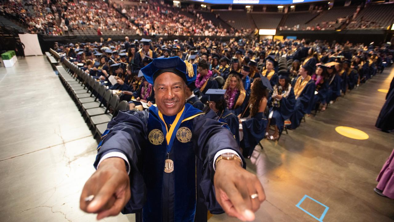 Chancellor May points his fingers at the camera and smiles with graduates from the class of 2025 celebrating behind him.