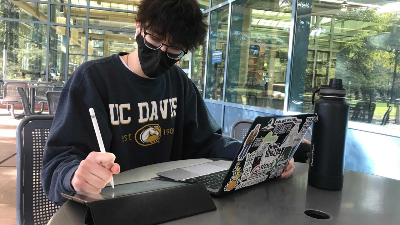 Student with mask working on laptop and tablet with UC Davis sweatshirt