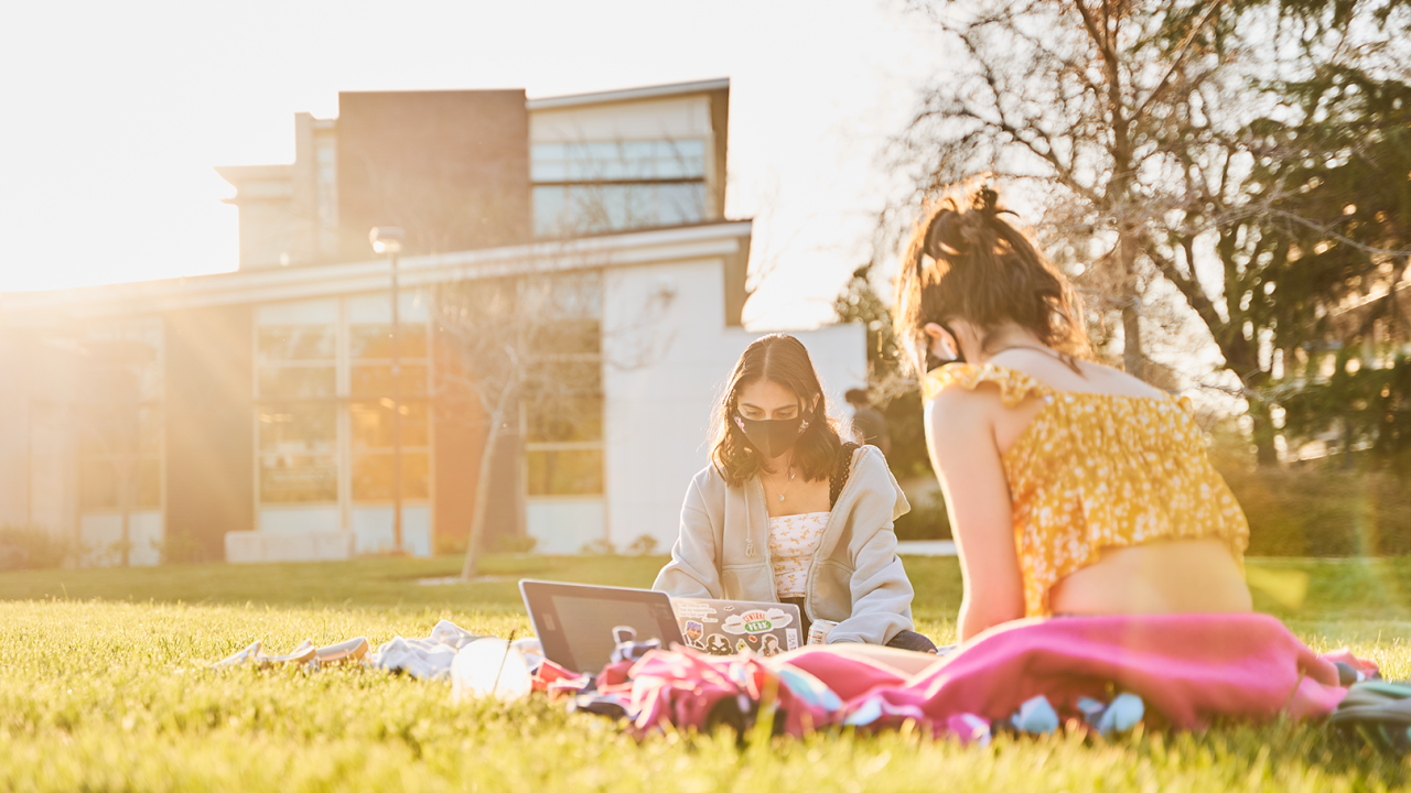 Two students wearing face masks sitting on picnic blanket outside of residence hall
