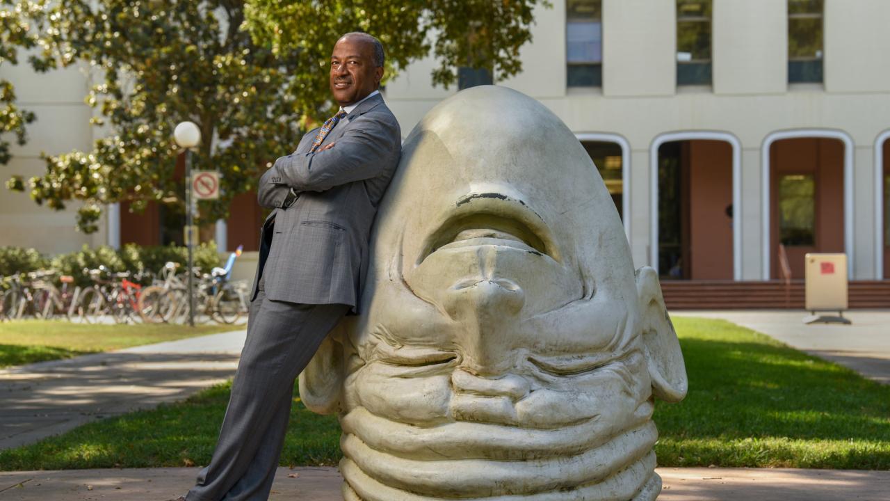 Chancellor Gary S. May, in suit, leans against "Eye on Mrak" Egghead sculpture.