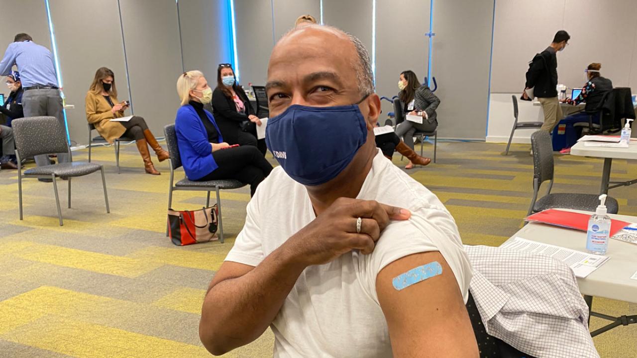 Chancellor Gary S. May, wearing a mask, rolls up T-shirt sleeve to show bandage over innoculation.