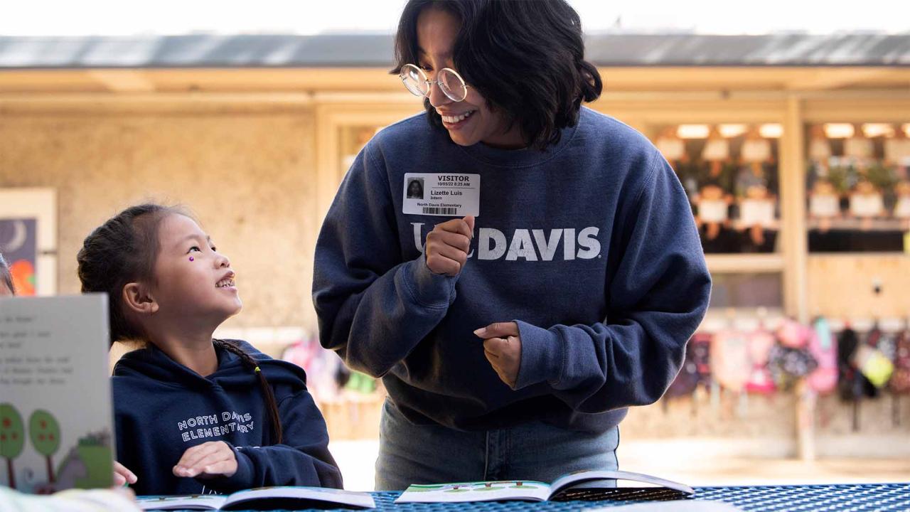 A college student smiling down at an elementary student reading a book.