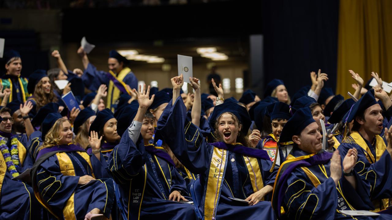 Graduates in navy and gold graduation robes cheering and tossing caps at commencement