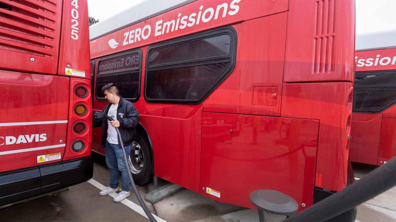 A male student dressed in a black jacket with blue jeans holds a black electric charging cord in his left hand to prepare a full charge for a parked red Unitrans bus.