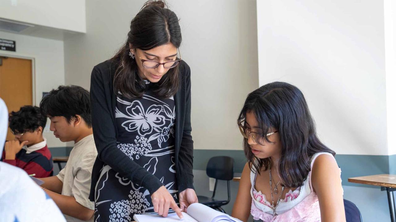 A teacher stands next to a seated student, pointing at a book on the desk, while other students work in the background.
