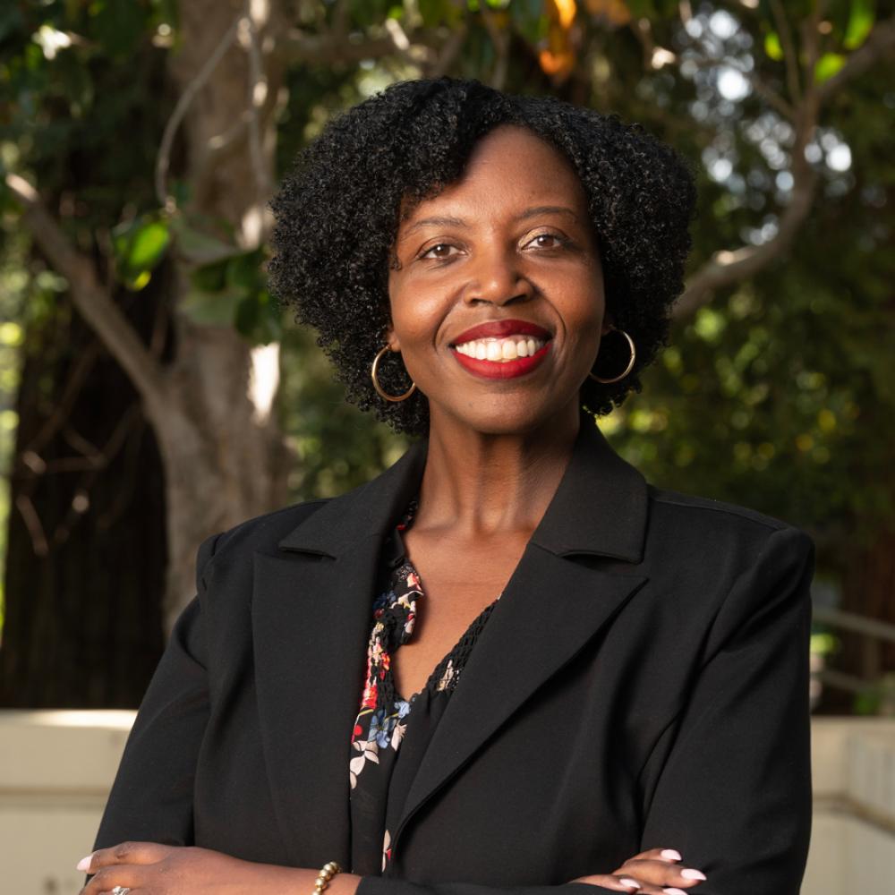 Smiling woman in a black blazer with arms crossed, standing outdoors among trees.