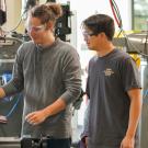 A male student wearing a grey long-sleeve shirt and protective clear goggles operates machinery in a student workshop, while a male student stands to the right to watch wearing a blue t-shirt and clear protective goggles.