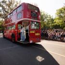 A red double-decker bus drives down the street in front of a parade crowd.