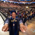 Chancellor May points his fingers at the camera and smiles with graduates from the class of 2025 celebrating behind him.
