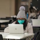 Student behind laptop with mask and hat.