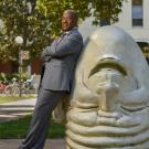 Chancellor Gary S. May, in suit, leans against "Eye on Mrak" Egghead sculpture.
