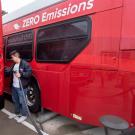 A male student dressed in a black jacket with blue jeans holds a black electric charging cord in his left hand to prepare a full charge for a parked red Unitrans bus. 