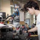 A student in a white T-shirt working on equipment in an engineering lab.
