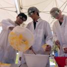 Students dressed in white lab coats and goggles mixing a yellow mixture of ice cream in a bowl under a white tent